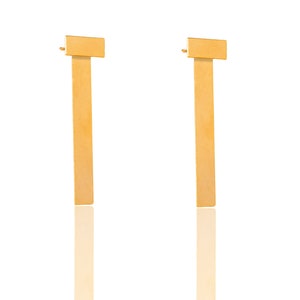 May include: A pair of gold-toned earrings with a long, rectangular shape. The earrings have a simple, minimalist design.