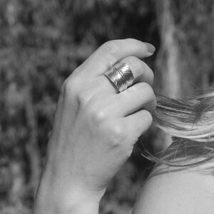 May include: A silver ring with a leaf design. The ring is worn on the middle finger of a person's hand.