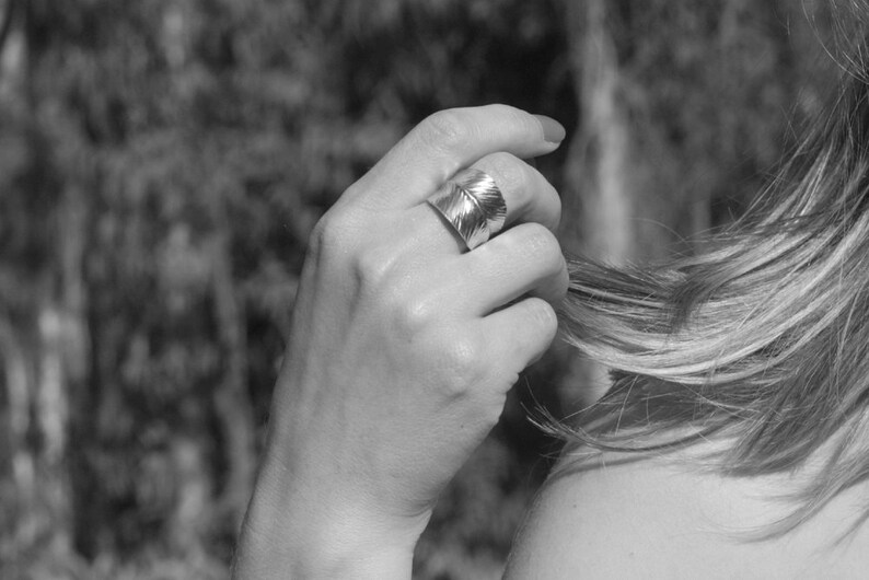 May include: A silver ring with a leaf design. The ring is worn on the middle finger of a person's hand.