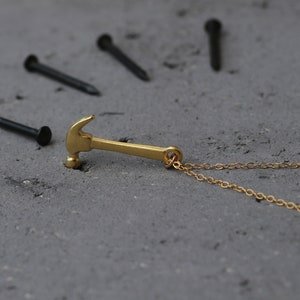 May include: A gold hammer pendant necklace with a delicate gold chain. The hammer is a simple design with a rounded head and a straight handle. The necklace is lying on a gray surface with several black nails scattered around it.
