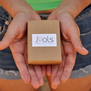 May include: A person is holding a small brown box with the words "Jools Wear your story" printed on it.