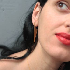 Könnte beinhalten: Eine Frau mit langen schwarzen Haaren trägt goldene Dreiecksohrringe. Sie trägt roten Lippenstift und hat einen neutralen Gesichtsausdruck.