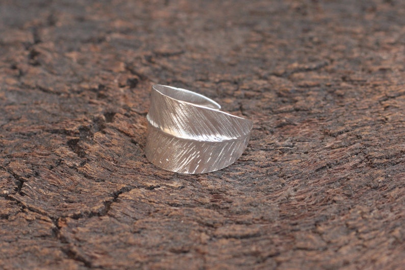 May include: A silver ring shaped like a feather, with a textured surface. The ring is on a brown wooden background.