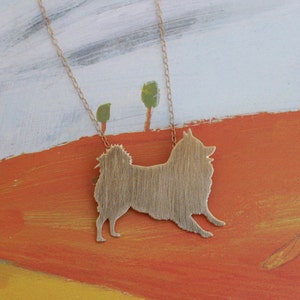 Op de afbeelding: Een gouden ketting met een hanger in de vorm van een silhouet van een hond van geborsteld metaal. De hanger hangt tegen een geschilderde achtergrond van een landschap met bomen en een heuvel.