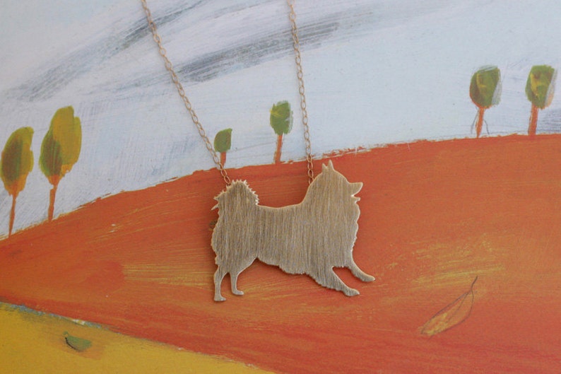 Op de afbeelding: Een gouden ketting met een hanger in de vorm van een silhouet van een hond van geborsteld metaal. De hanger hangt tegen een geschilderde achtergrond van een landschap met bomen en een heuvel.