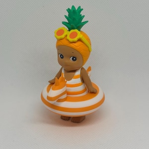 Op de afbeelding: Een klein figuurtje met een ananasvormige hoed met groene bladeren en een gele zonnebril. Het figuurtje draagt een wit met oranje gestreept badpak en bevindt zich in een oranje met wit gestreepte opblaasbare zwemband.