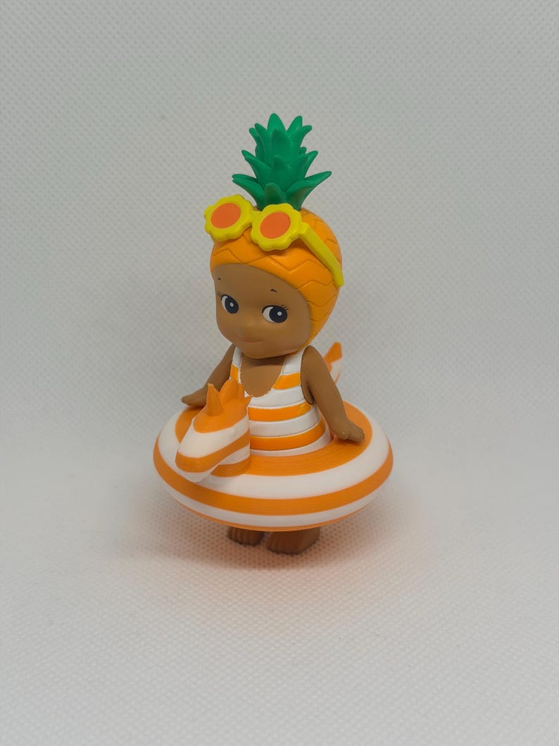 Op de afbeelding: Een klein figuurtje met een ananasvormige hoed met groene bladeren en een gele zonnebril. Het figuurtje draagt een wit met oranje gestreept badpak en bevindt zich in een oranje met wit gestreepte opblaasbare zwemband.