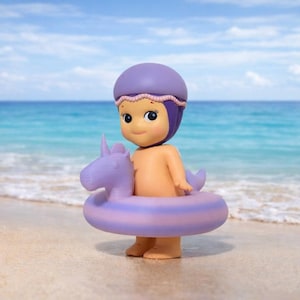 Puede incluir: Una pequeña figura desnuda con gorro de baño morado y flotador con forma de unicornio en una playa de arena. La figura tiene una cara sonriente, con el océano y el cielo azul de fondo.