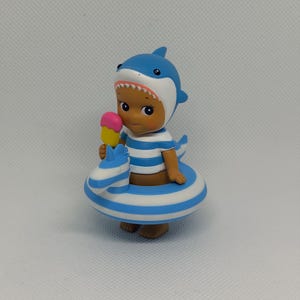 Op de afbeelding: Een klein kinderfiguurtje met een blauw-witte haaienkap, een blauw-wit gestreept shirt en een blauw-wit gestreepte opblaasbare zwemband in de vorm van een zeepaardje. Het figuurtje houdt een geel ijsje met een roze bovenkant vast.