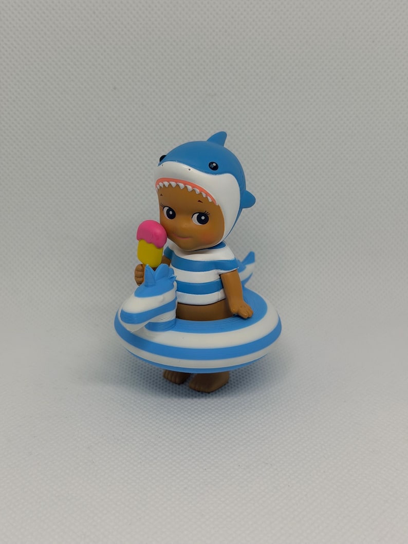 Op de afbeelding: Een klein kinderfiguurtje met een blauw-witte haaienkap, een blauw-wit gestreept shirt en een blauw-wit gestreepte opblaasbare zwemband in de vorm van een zeepaardje. Het figuurtje houdt een geel ijsje met een roze bovenkant vast.