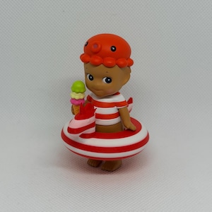Op de afbeelding: Een klein figuurtje met een oranje octopusmuts en een rood-wit gestreept shirt. Het figuurtje zit in een rood-wit gestreepte opblaasbare zwemband in de vorm van een zeepaardje en houdt een klein ijshoorntje met drie bolletjes ijs vast.