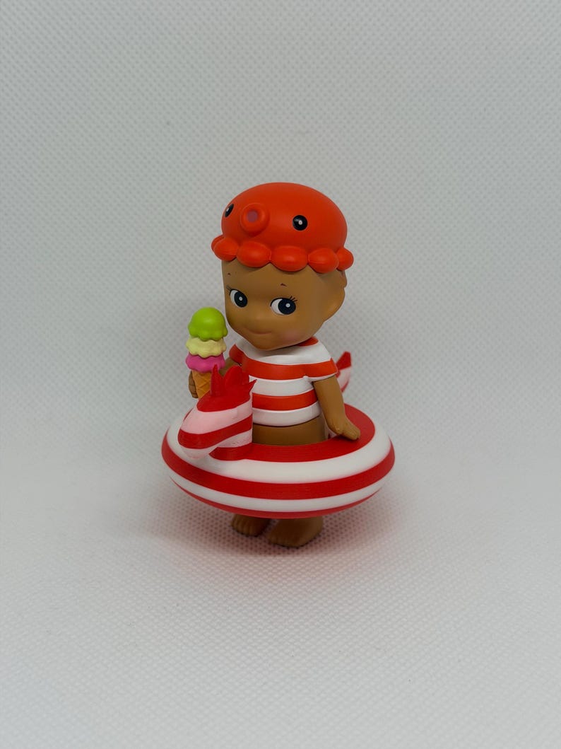 Op de afbeelding: Een klein figuurtje met een oranje octopusmuts en een rood-wit gestreept shirt. Het figuurtje zit in een rood-wit gestreepte opblaasbare zwemband in de vorm van een zeepaardje en houdt een klein ijshoorntje met drie bolletjes ijs vast.