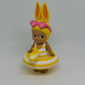 Op de afbeelding: Een klein beeldje van een figuurtje met gele konijnenoren en een roze zonnebril in de vorm van hartjes. Het figuurtje draagt een geel-wit gestreept badpak en zit in een bijpassende geel-wit gestreepte opblaasbare zwemband.