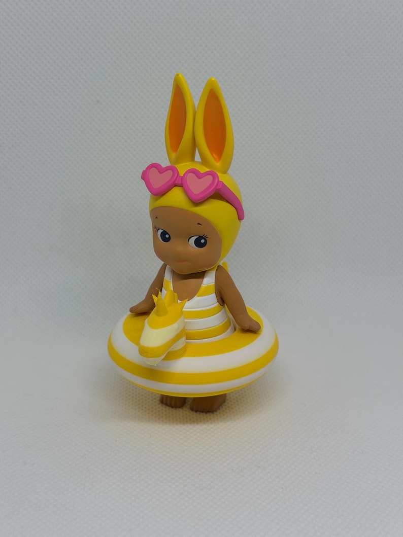 Op de afbeelding: Een klein beeldje van een figuurtje met gele konijnenoren en een roze zonnebril in de vorm van hartjes. Het figuurtje draagt een geel-wit gestreept badpak en zit in een bijpassende geel-wit gestreepte opblaasbare zwemband.