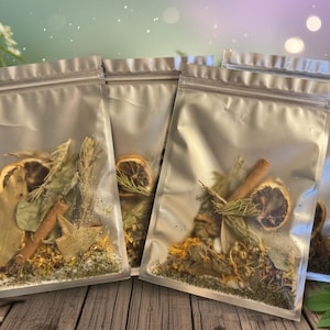 May include: Four clear, resealable bags filled with dried herbs, spices, and citrus slices. The bags contain cinnamon sticks, dried orange slices, and various green herbs. The bags are arranged on a wooden surface with a blurred background.