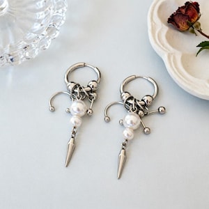 May include: A pair of silver-toned hoop earrings with a unique design. Each earring features a cluster of small beads, three dangling pearls, and a pointed, dagger-like charm. The earrings are displayed on a light blue surface.