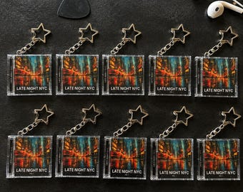 May include: Ten square keychains featuring a miniature CD case design with a city street scene at night. Each keychain has a star-shaped metal ring and the text LATE NIGHT NYC VOL 1 printed on the front. A black guitar pick is included.
