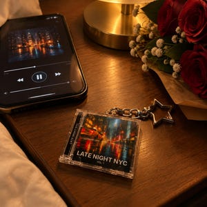 May include: A clear plastic keychain shaped like a miniature CD jewel case featuring a city street scene at night with the text LATE NIGHT NYC VOL. 1 printed on the front, attached to a silver-toned star-shaped metal keyring.