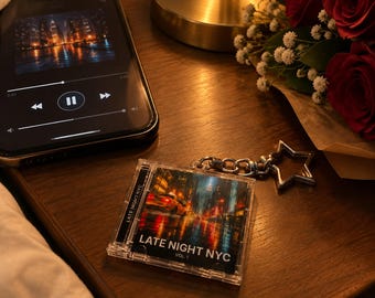May include: A clear plastic keychain shaped like a miniature CD jewel case featuring a city street scene at night with the text LATE NIGHT NYC VOL. 1 printed on the front, attached to a silver-toned star-shaped metal keyring.
