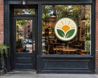 May include: A circular window decal featuring a stylized orange sun rising above two green leaves, displayed on the glass storefront of a building with a black door and frame, set against a red brick exterior wall with visible interior seating.