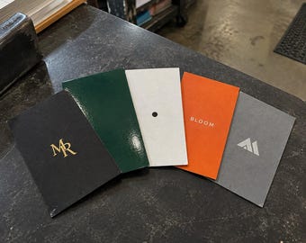 May include: Five rectangular cards arranged in a fan shape. The first is black with gold MR text, the second is dark green, the third is white with a black dot, the fourth is orange with the word BLOOM, and the fifth is gray with a white logo.