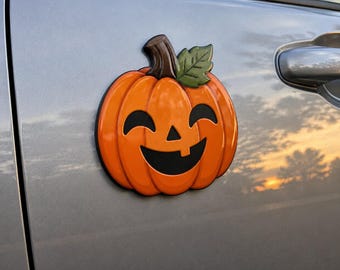 May include: A glossy orange pumpkin emblem with a brown stem and a single green leaf. The pumpkin features a black jack-o'-lantern face with two curved eyes, a triangular nose, and a wide, toothy grin, attached to a metallic silver car door.