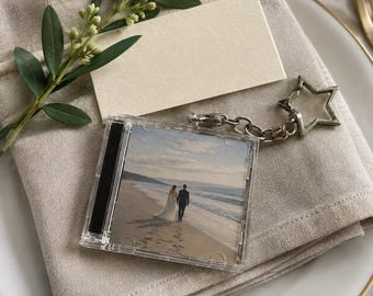 May include: A square clear plastic keychain featuring a miniature CD case design. The insert displays a photograph of two people walking along a sandy beach near the ocean. The keychain includes a silver-toned metal star-shaped clasp.