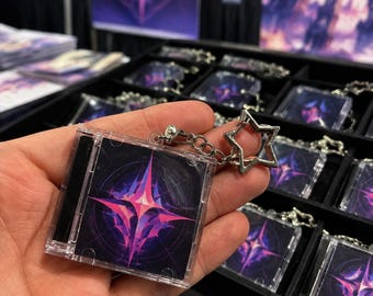 May include: A square keychain designed as a miniature CD jewel case featuring a dark purple and black background with a vibrant, glowing pink and blue four-pointed star graphic. It includes a silver-toned metal star-shaped clasp.