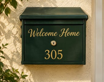May include: A dark green metal mailbox with a slanted top and a central key lock. The front panel features the gold text "Welcome Home" in a cursive script above the gold numbers "305" in a clean, sans-serif font, mounted on a textured wall.