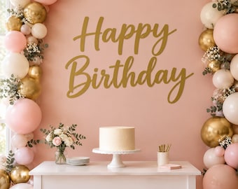 May include: A gold script sign reading Happy Birthday mounted on a peach wall, flanked by balloon arches in shades of pink, cream, and metallic gold. A white table holds a plain cake on a stand, a small vase of flowers, and a stack of plates.