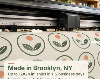 May include: A large-format printer head positioned over a roll of off-white vinyl material featuring a repeating pattern of dark green circular outlines, each containing two dark green leaves and a small solid orange circle above them.