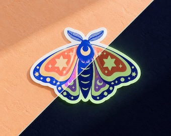 May include: A moth sticker featuring a vibrant design with a crescent moon on its head, star patterns on its wings, and a color palette of blue, orange, purple, and green. The sticker displays a split effect showing a glow-in-the-dark feature.