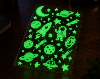 May include: A rectangular sheet of glow-in-the-dark stickers featuring various space-themed designs including a crescent moon, planets with rings, a rocket ship, an astronaut, a flying saucer, a spiral galaxy, comets, and numerous stars.