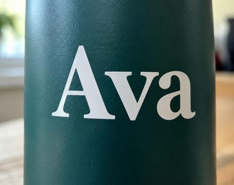 May include: A dark forest green cylindrical water bottle featuring the name Ava printed in a bold, white, serif-style font across the center. The surface of the bottle has a smooth, matte finish, providing a clean and simple aesthetic.
