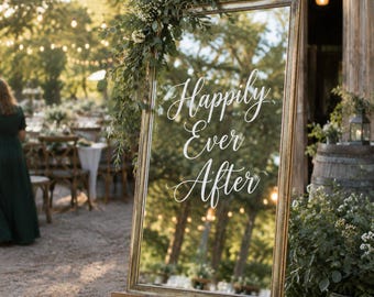 May include: A tall, rectangular mirror with a metallic gold frame, mounted on a wooden easel. The glass surface features the white script text "Happily Ever After" and is decorated with a lush arrangement of green foliage and small white flowers.
