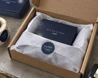 May include: A brown cardboard shipping box containing a stack of dark blue business cards with the text RIVER & OAK and a wave icon. The contents are wrapped in white tissue paper secured with a round dark blue sticker that says THANK YOU.