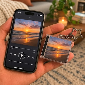 Puede incluir: Un teléfono inteligente que muestra una interfaz de reproductor de música con una imagen de playa al atardecer y un llavero miniatura de caja de CD a juego con la misma escena, unido a una anilla metálica plateada en forma de estrella.