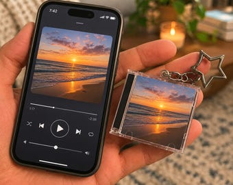 May include: A smartphone displaying a music player interface with a sunset beach image and a matching miniature CD case keychain featuring the same sunset beach scene, attached to a silver-toned star-shaped metal keyring.