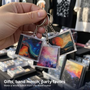 May include: Three square keychains featuring miniature CD jewel case designs. Each keychain displays a unique colorful graphic print, including abstract blue and orange swirls, a pink and purple sky, and a sunset landscape with a large orange moon.