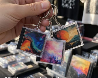 May include: Three square keychains featuring miniature CD jewel case designs. Each keychain displays a unique colorful graphic print, including abstract blue and orange swirls, a pink and purple sky, and a sunset landscape with a large orange moon.