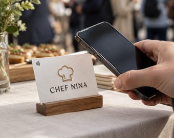 May include: A white rectangular NFC payment card featuring a gold chef hat icon and the text CHEF NINA in black lettering. The card is held upright in a dark brown wooden stand, while a black smartphone is positioned nearby for contactless payment.