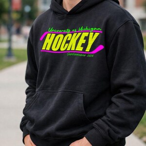 May include: Black hooded sweatshirt with a front pocket. The sweatshirt features the text "University of Michigan HOCKEY Sophomore 2026" in neon green and yellow with a pink hockey stick graphic.