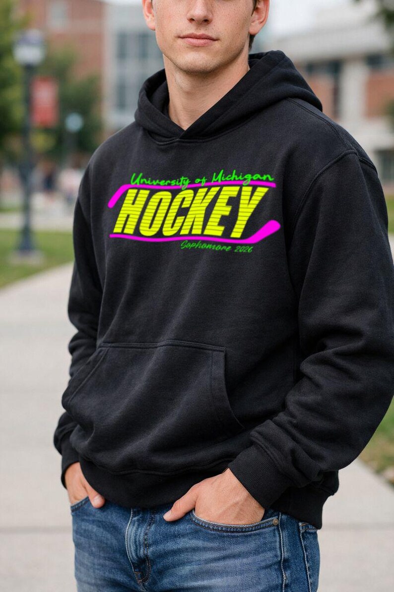 May include: Black hooded sweatshirt with a front pocket. The sweatshirt features the text "University of Michigan HOCKEY Sophomore 2026" in neon green and yellow with a pink hockey stick graphic.