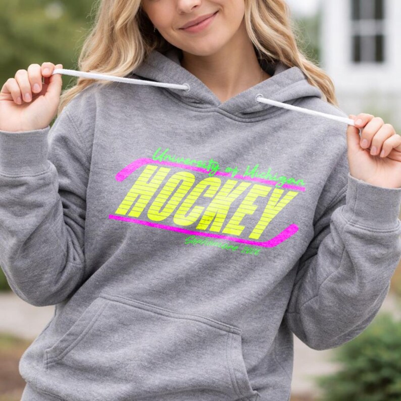 May include: A gray hoodie with a front pocket and a drawstring hood. The hoodie features the text "University of Michigan HOCKEY" in neon yellow and pink. The text is in a stylized font.