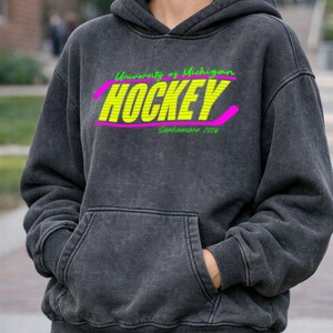 May include: Dark gray hoodie with a front pocket and a hood. The front features the text "University of Michigan HOCKEY Sophomore 2026" in neon green and yellow, with a pink hockey stick graphic.