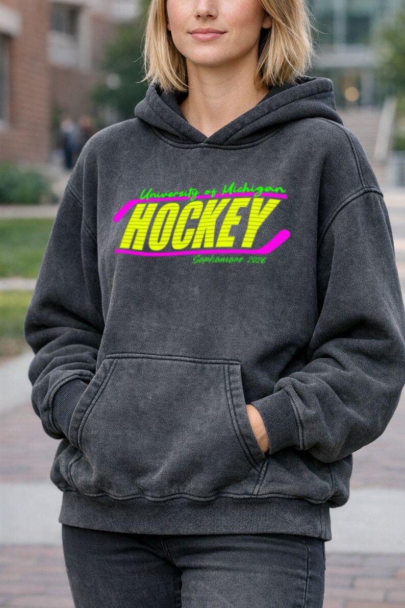 May include: Dark gray hoodie with a front pocket and a hood. The front features the text "University of Michigan HOCKEY Sophomore 2026" in neon green and yellow, with a pink hockey stick graphic.