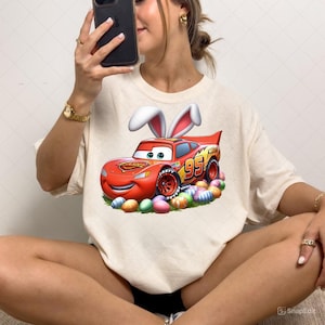 May include: Cream-colored t-shirt featuring a cartoon image of Lightning McQueen with bunny ears and the number 95. The design includes colorful Easter eggs. The shirt is a fun, festive item for Easter.