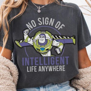 May include: Dark gray t-shirt featuring a graphic of Buzz Lightyear with the text "NO SIGN OF INTELLIGENT LIFE ANYWHERE". The cartoon character is in a flying pose, with green, purple, and white accents. The text is in white and purple.