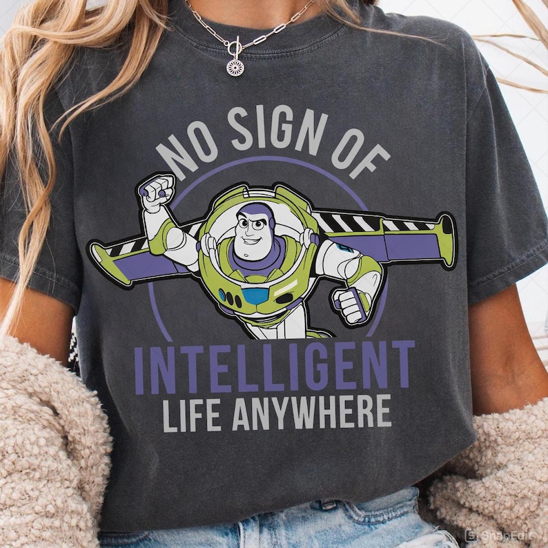 May include: Dark gray t-shirt featuring a graphic of Buzz Lightyear with the text "NO SIGN OF INTELLIGENT LIFE ANYWHERE". The cartoon character is in a flying pose, with green, purple, and white accents. The text is in white and purple.