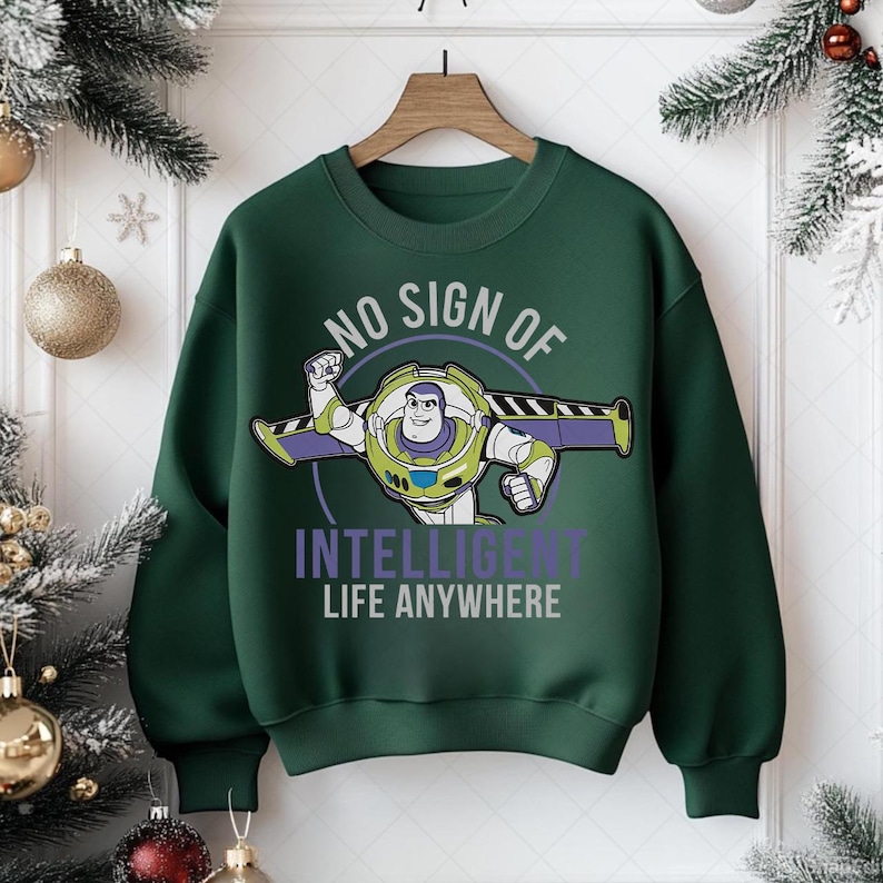 May include: A dark green sweatshirt featuring a graphic of Buzz Lightyear with the text "NO SIGN OF INTELLIGENT LIFE ANYWHERE". The design includes Buzz in his space suit with outstretched arms. The sweatshirt is hanging on a wooden hanger.